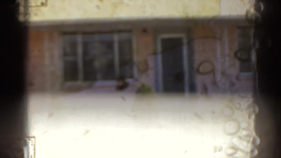 1962 Florida home-movie frame showing a pink house facade with a blue door, windows, and small shrubs, softened by film wear.