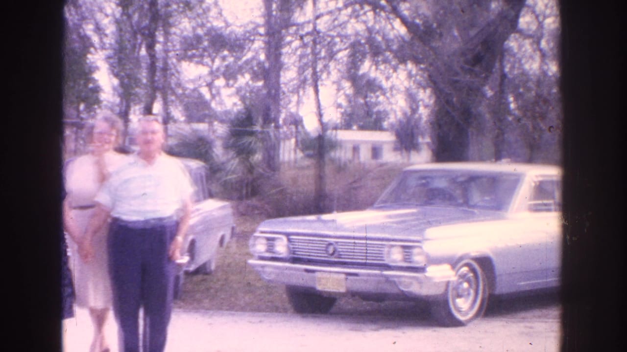 1962 Florida home-movie frame showing three adults posed beside a light-colored sedan near trees and residential houses.