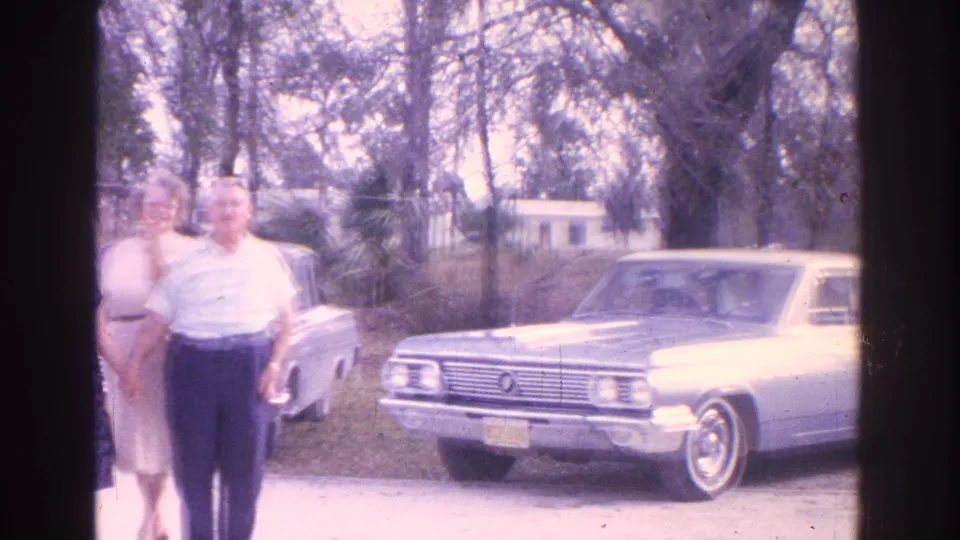 1962 Florida home-movie frame showing three adults posed beside a light-colored sedan near trees and residential houses.