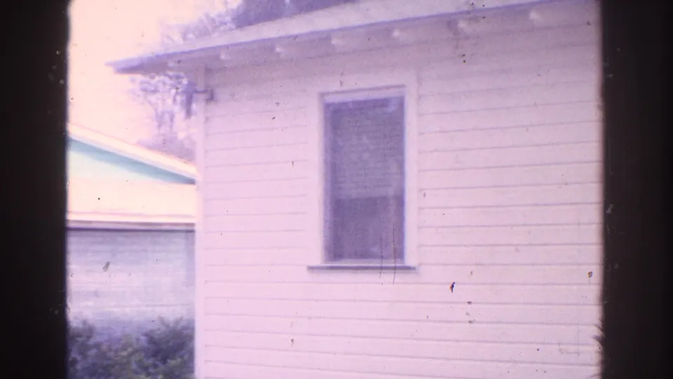 1962 Florida home-movie frame centered on a white clapboard house wall with a window and blue outbuilding at the edge.