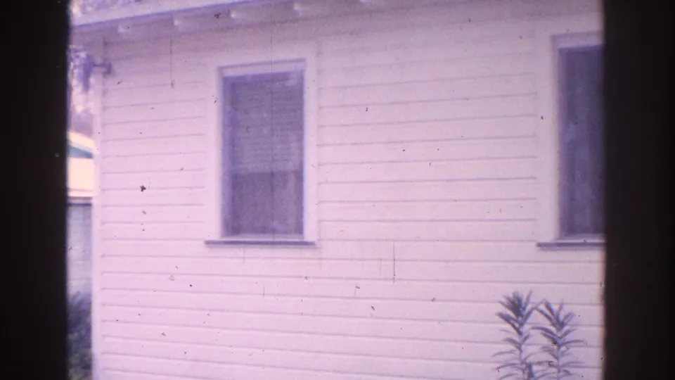 1962 Florida residential frame showing the side wall of a white house, two windows, and a small plant in the yard.