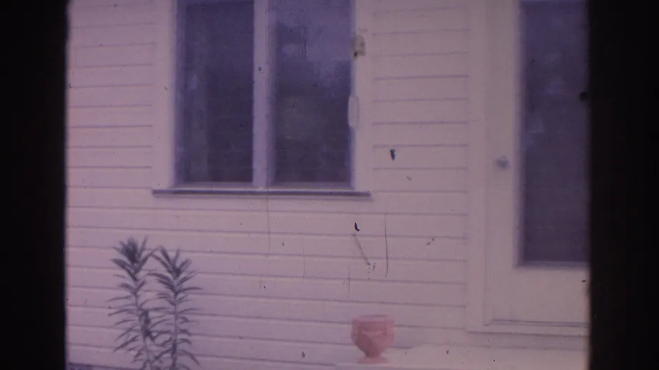 1962 Florida home-movie frame showing a white house exterior with paired windows, a door edge, and a small potted plant near the wall.