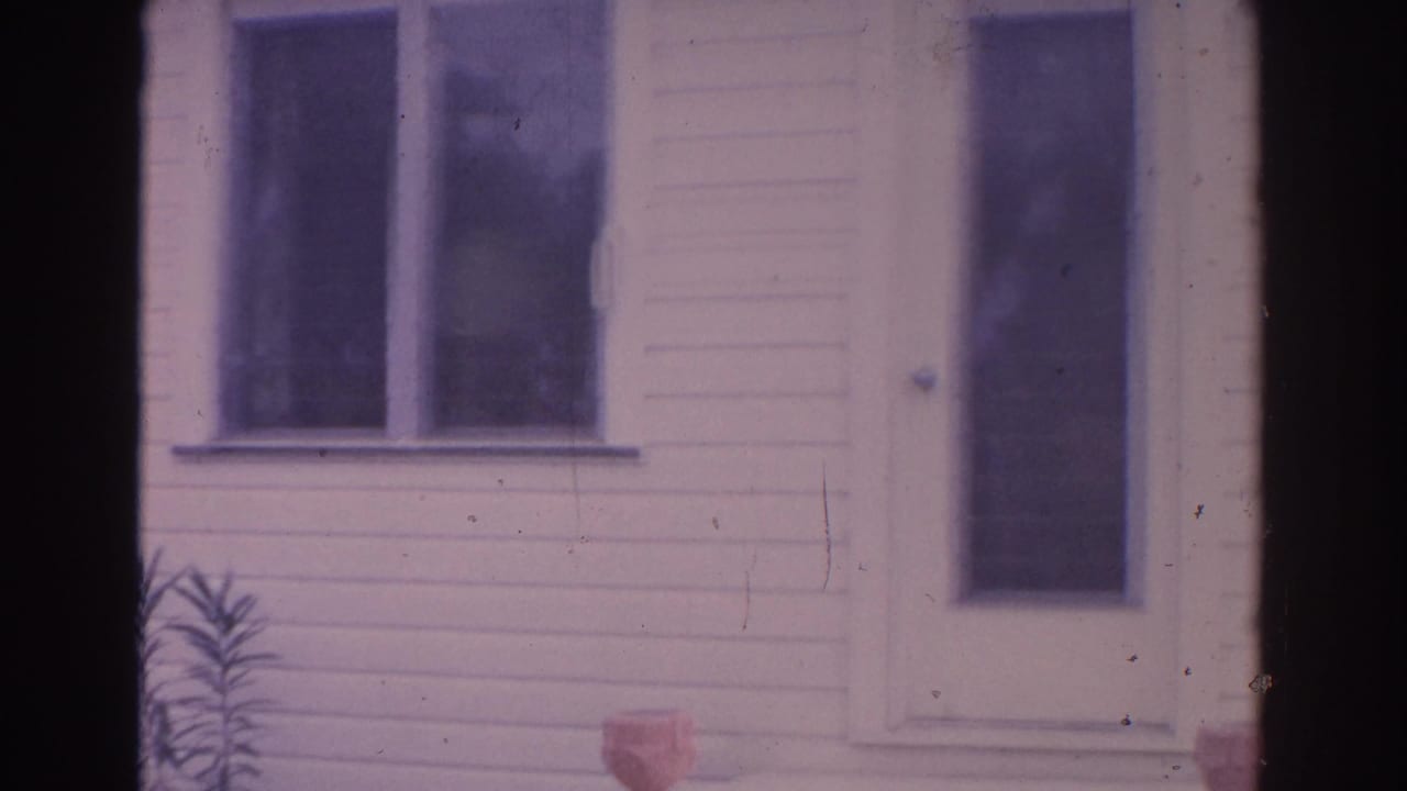 1962 Florida 8mm frame showing a white clapboard house facade with a front door, windows, porch urns, and a small plant at left.