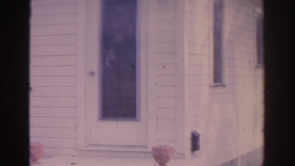 1962 Florida 8mm frame showing the front door area of a white clapboard house, with a tall window, porch urn, and side wall visible.