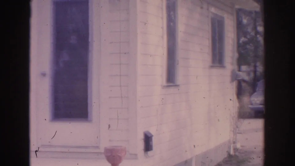 1962 Florida home-movie frame showing the house corner with front door, side windows, porch urn, wall-mounted box, and trees at right.