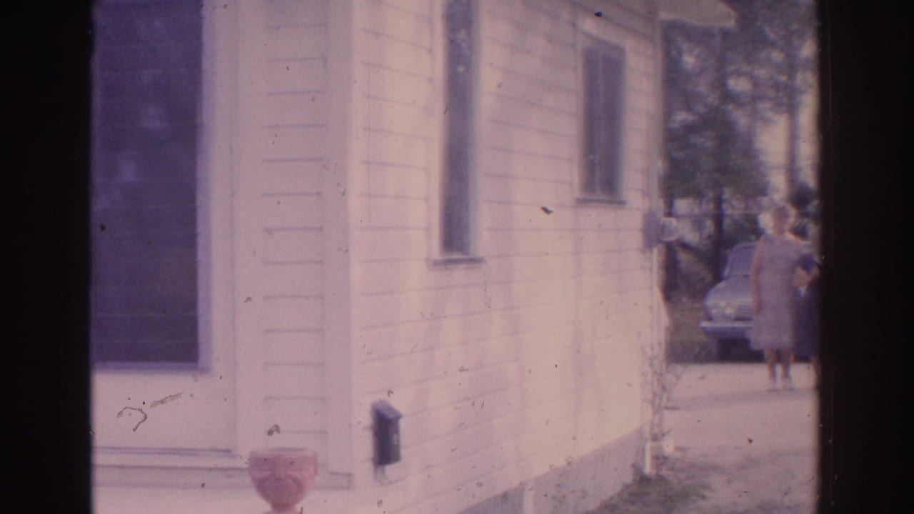 White-sided house beside a driveway with adults near a parked car in the background.