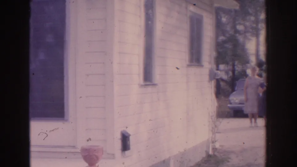 1962 Florida home-movie frame showing a light-sided house, driveway, parked car, and adults gathering near the far side.