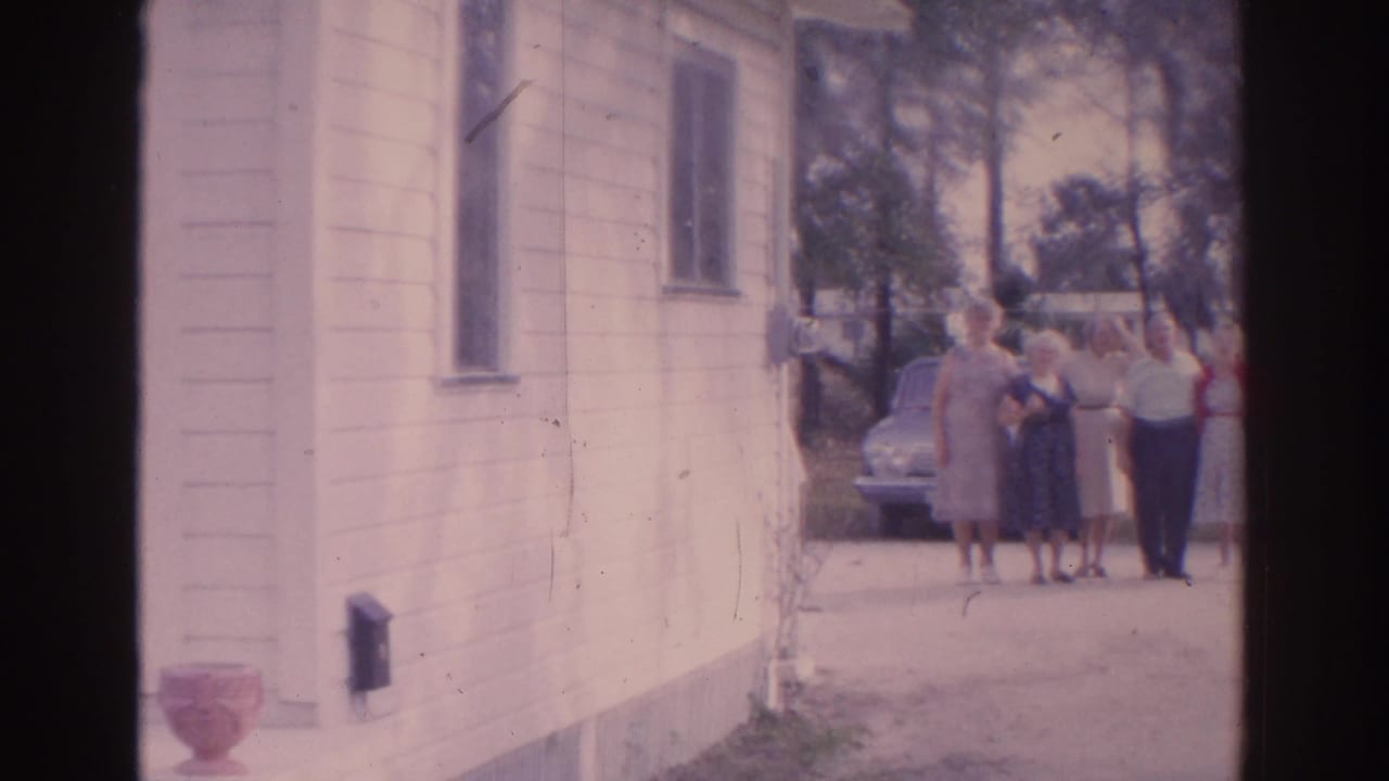 Adults stand near a parked car beside a light-sided house and trees.