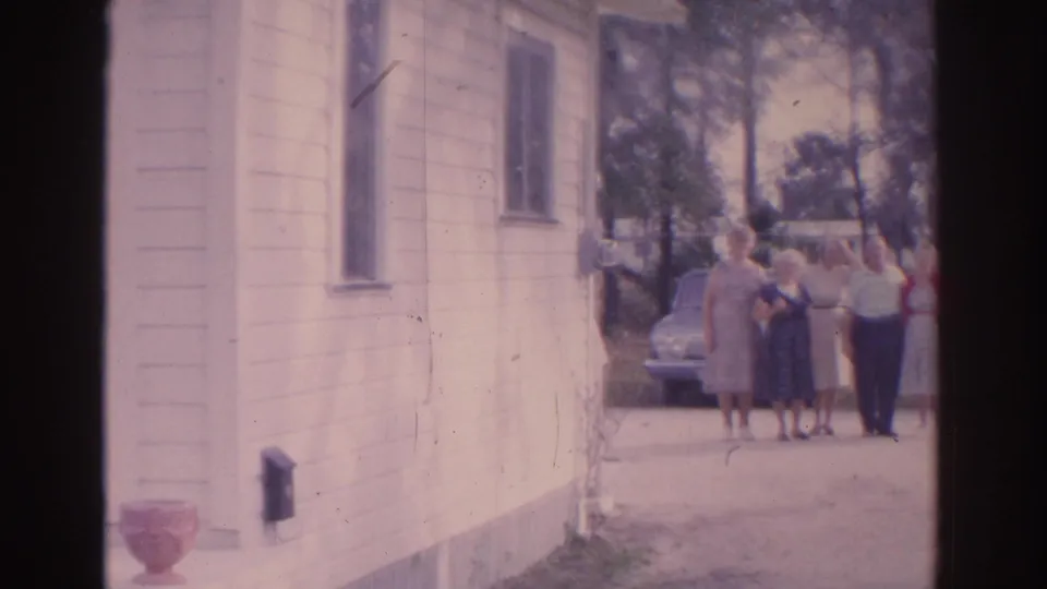 Adults in period clothing gather by a parked sedan beside a light-colored house in Florida home-movie footage from 1962.