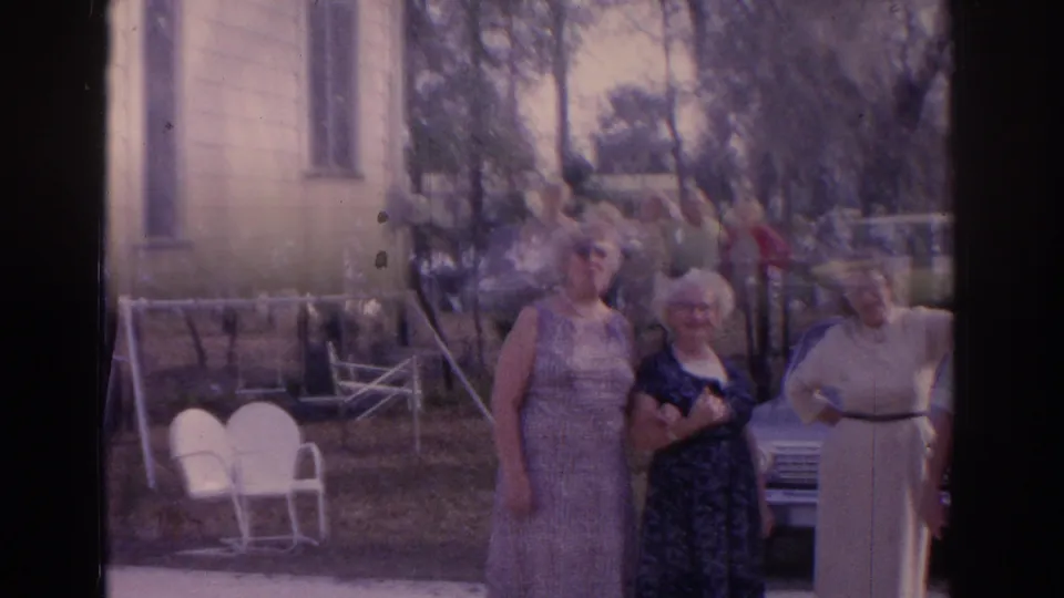 1962 Florida backyard frame showing adults posing together, with lawn chairs, trees, and a parked car behind them.