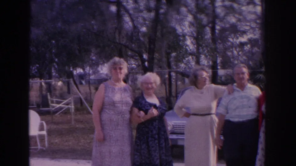 Four adults face the camera in a 1962 Florida backyard scene, with outdoor chairs, trees, and a parked car visible behind them.