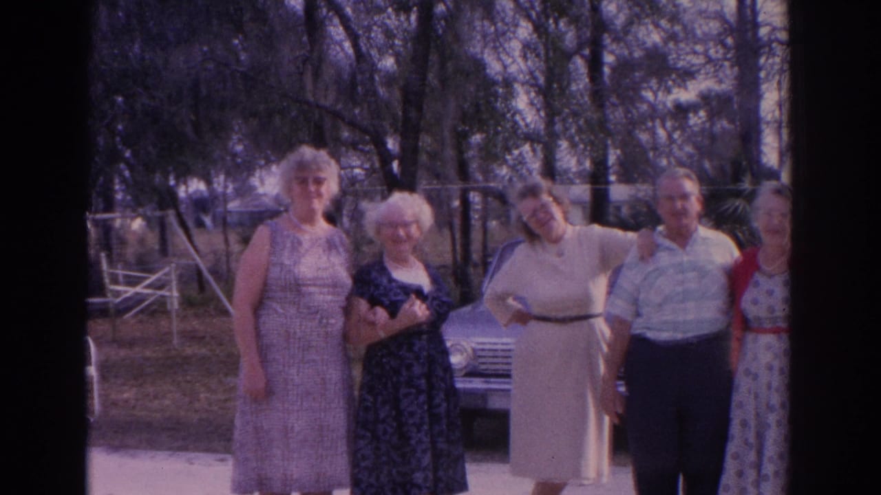 Five adults stand outdoors near a light-colored sedan in a tree-lined yard.