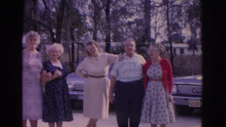 Well-dressed adults continue posing for an 8mm home-movie camera, with a light-colored car and trees in the background.