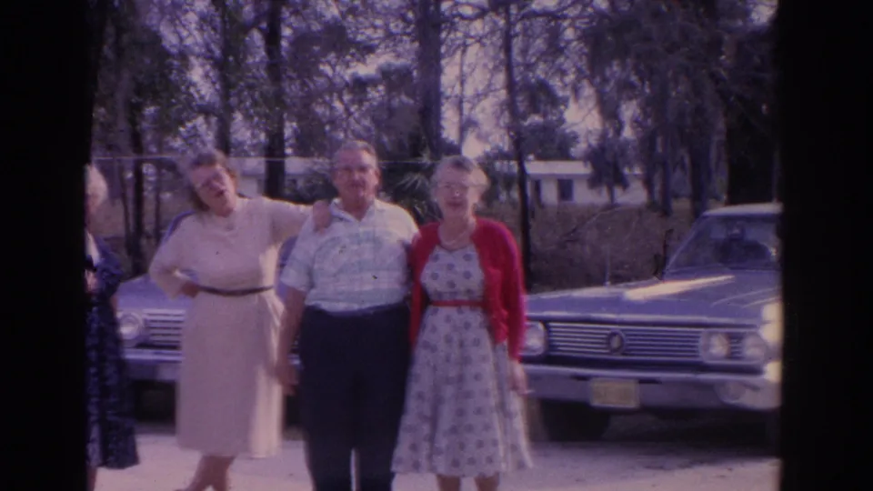 Final frame centers three adults in dress clothes beside the sedan, with another adult partly visible at the edge of the 8mm frame.
