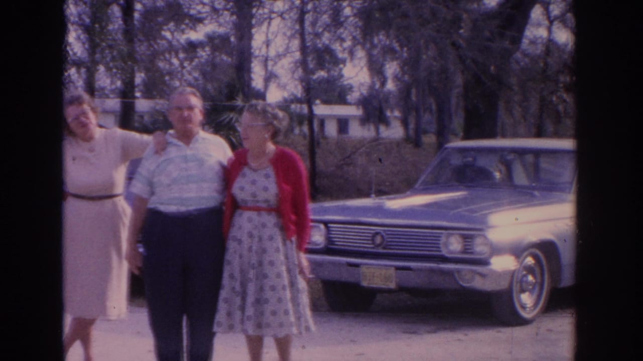 Three adults stand outdoors beside a blue car on a sunny day.