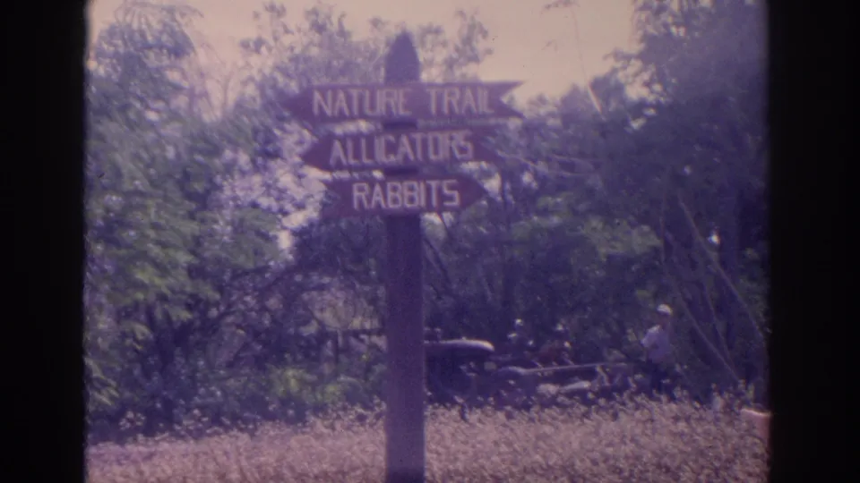 1962 Florida 8mm frame with the nature trail, alligators, and rabbits sign, plus a distant person and older vehicle in the background.