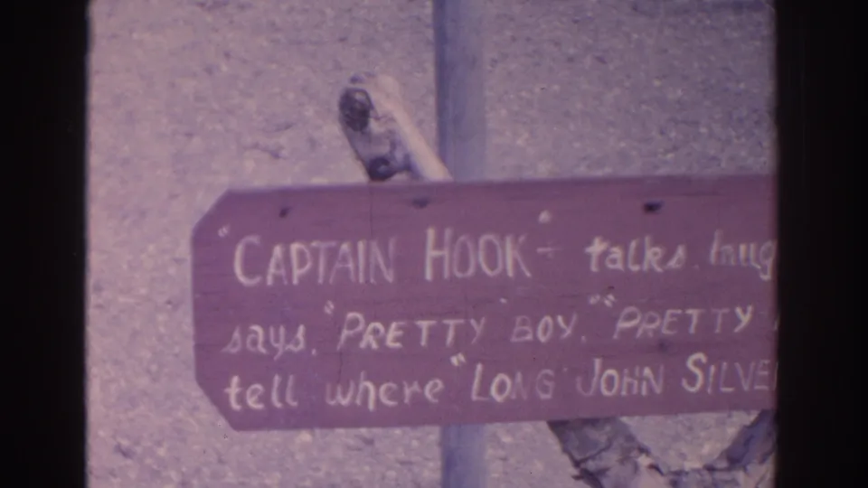 1962 Florida home-movie frame showing a reddish wooden attraction sign with handwritten references to Captain Hook and Long John Silver.