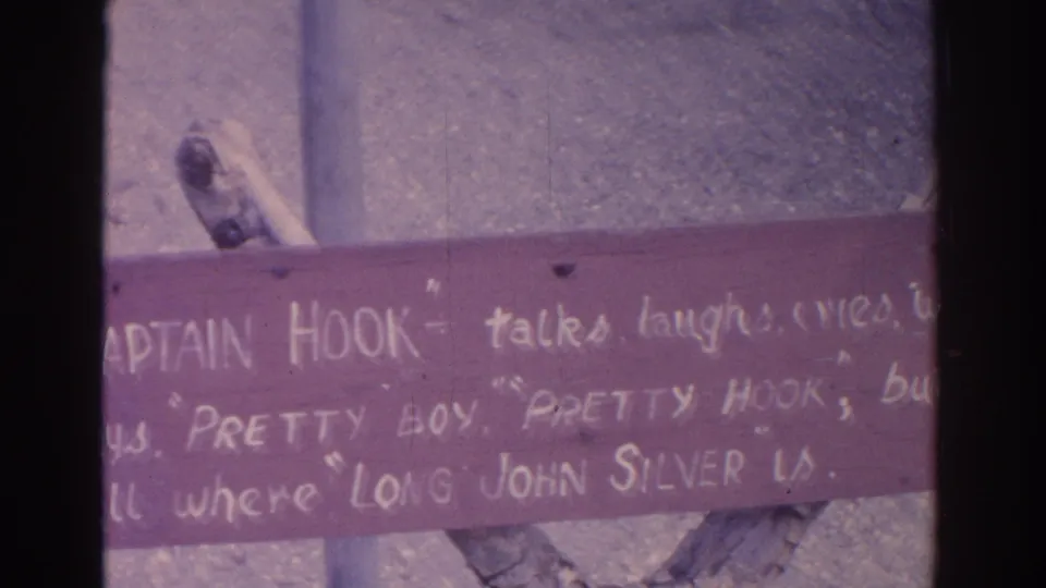 Partially cropped 1962 Florida home-movie frame of the themed wooden sign, with white lettering and a metal post visible.