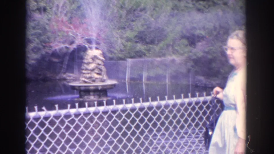 1962 Florida 8mm frame showing an adult woman in glasses near a chain-link fence, with a decorative fountain and pond behind her.