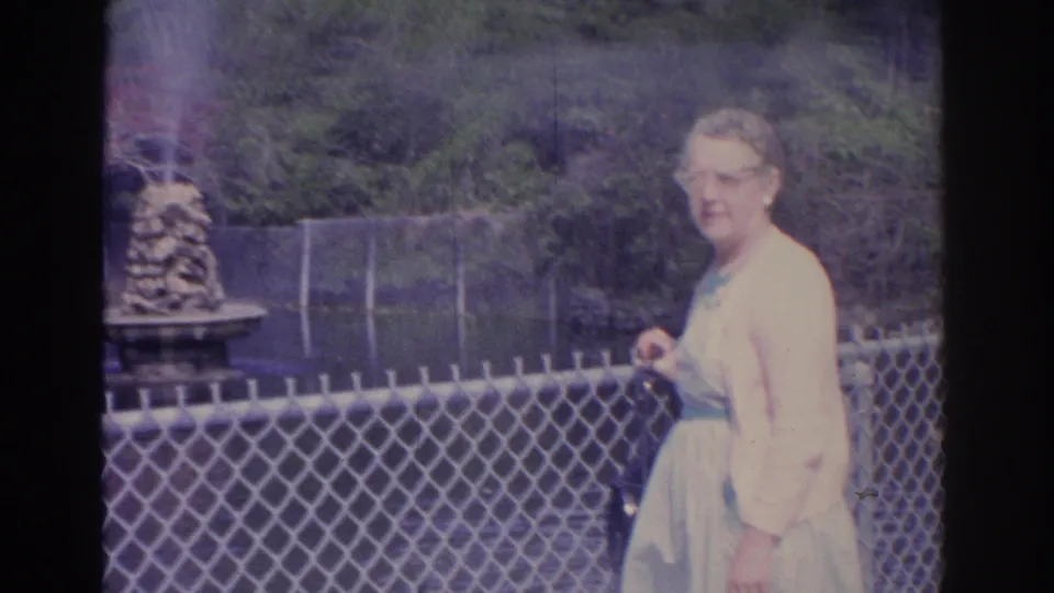 1962 Florida 8mm frame of an adult woman in glasses beside a chain-link fence, with the pond, fountain, and greenery in the background.