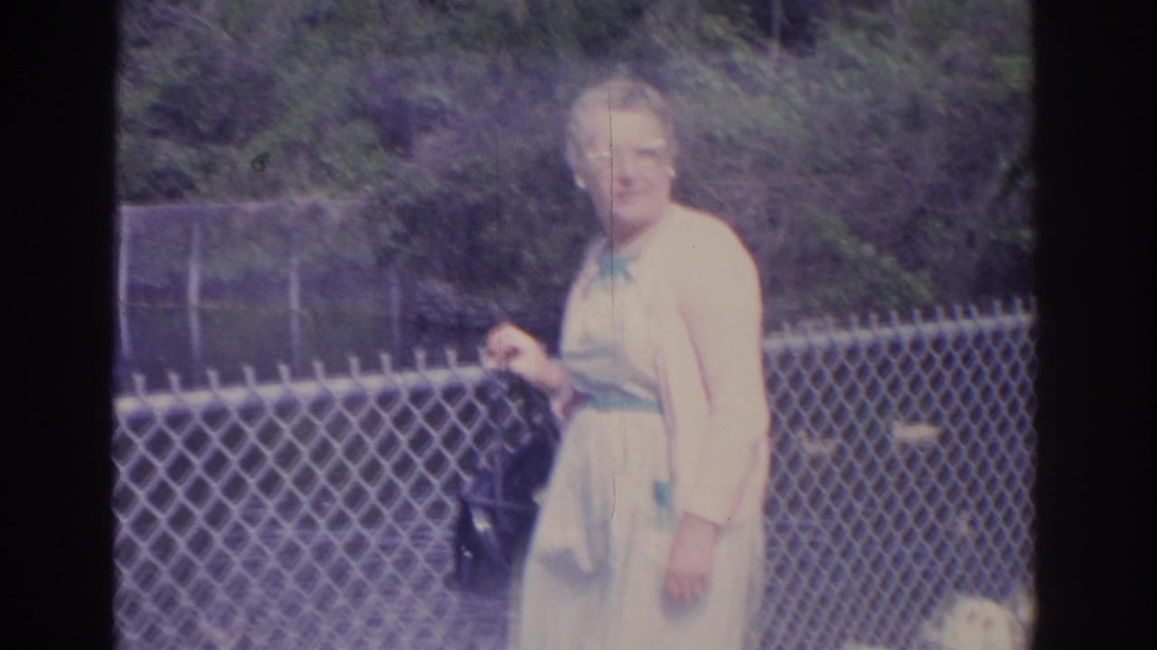 Older woman in a light dress and cardigan standing near a chain-link fence.