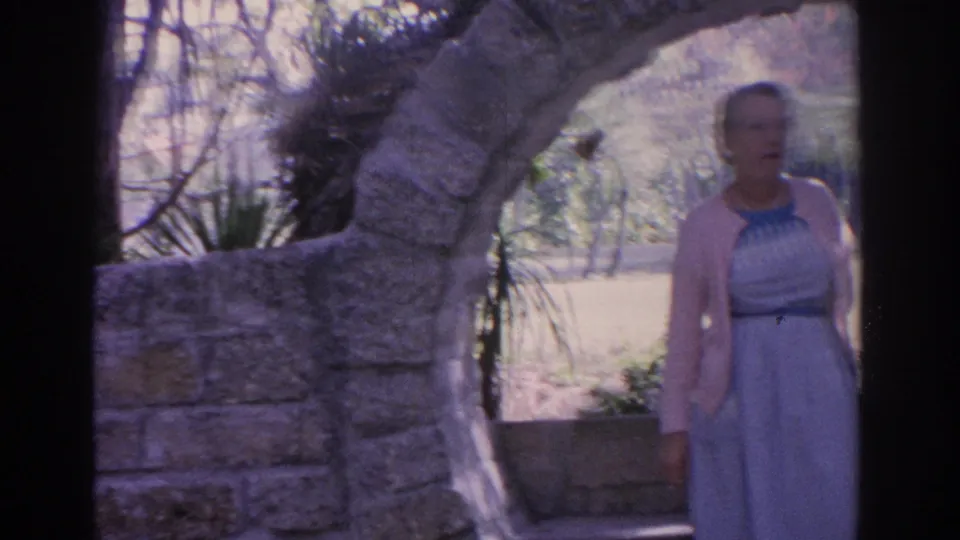 Older woman in a light dress and cardigan walks through a stone garden archway in 1962 Florida home-movie footage.