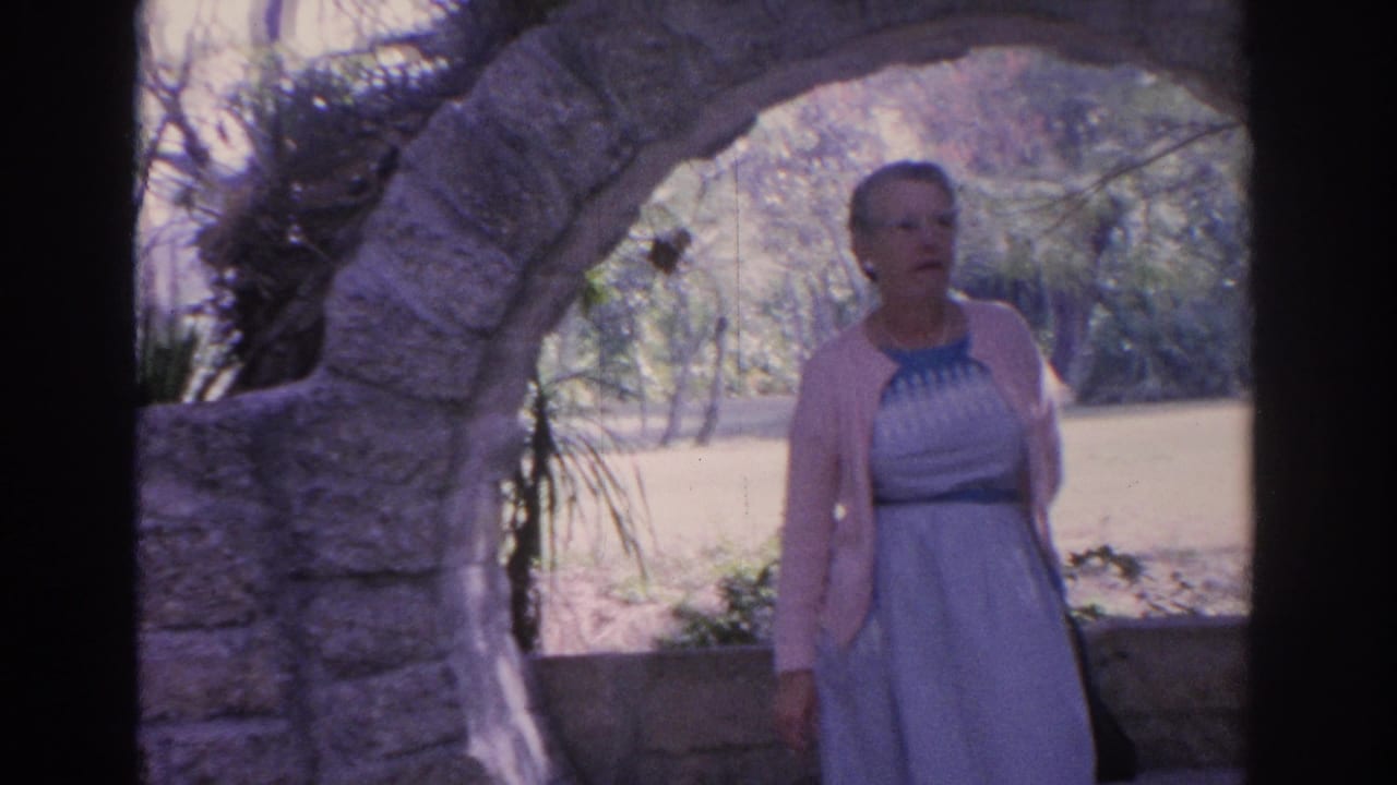 1962 Florida home-movie frame of a mature woman standing under a rustic stone arch with trees and sunlit ground behind her.