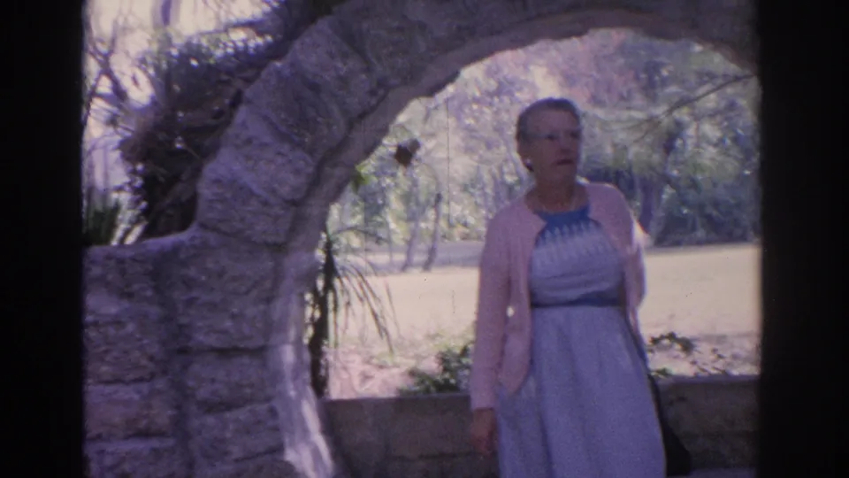 1962 Florida home-movie frame of a mature woman standing under a rustic stone arch with trees and sunlit ground behind her.