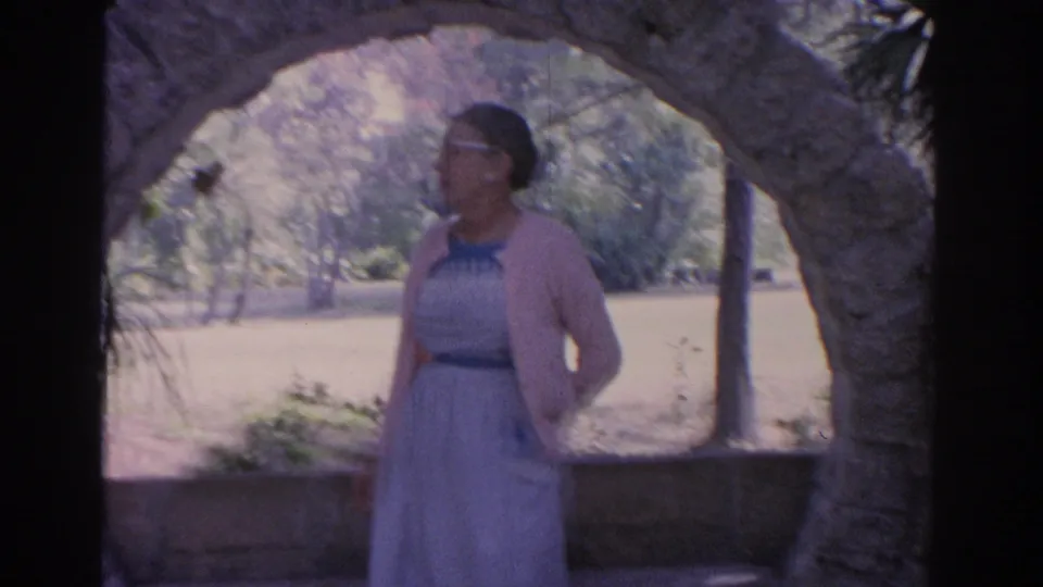 1962 Florida home-movie frame showing a mature woman in a cardigan looking sideways near a stone arch and shaded greenery.