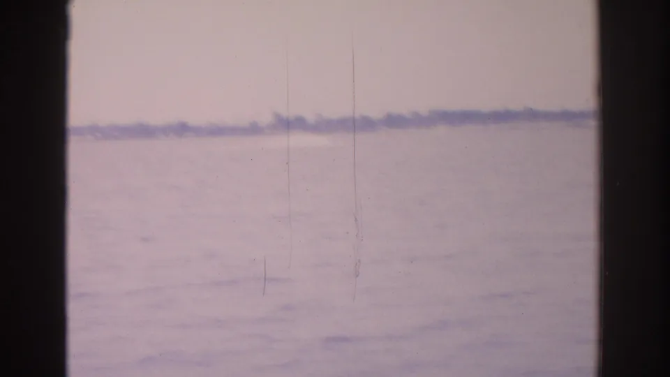 1962 Florida 8mm frame showing pale rippling water, hazy sky, and a low distant shoreline through dark gate edges.