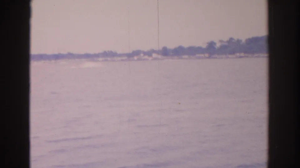 1962 Florida 8mm frame looking across calm water toward a distant tree-lined shore with scattered low houses.