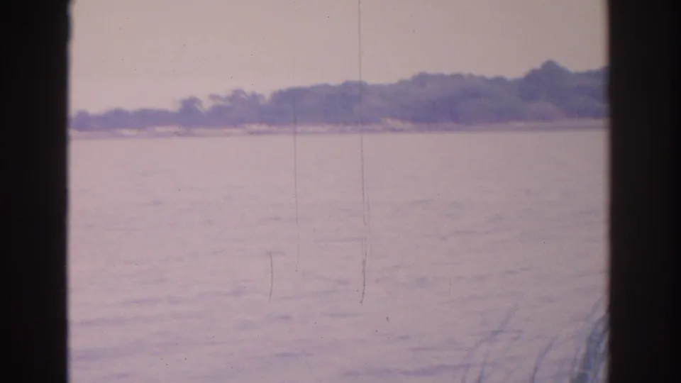 A soft 1962 Florida home-movie frame shows water in the foreground and a low, wooded shoreline with small houses beyond.