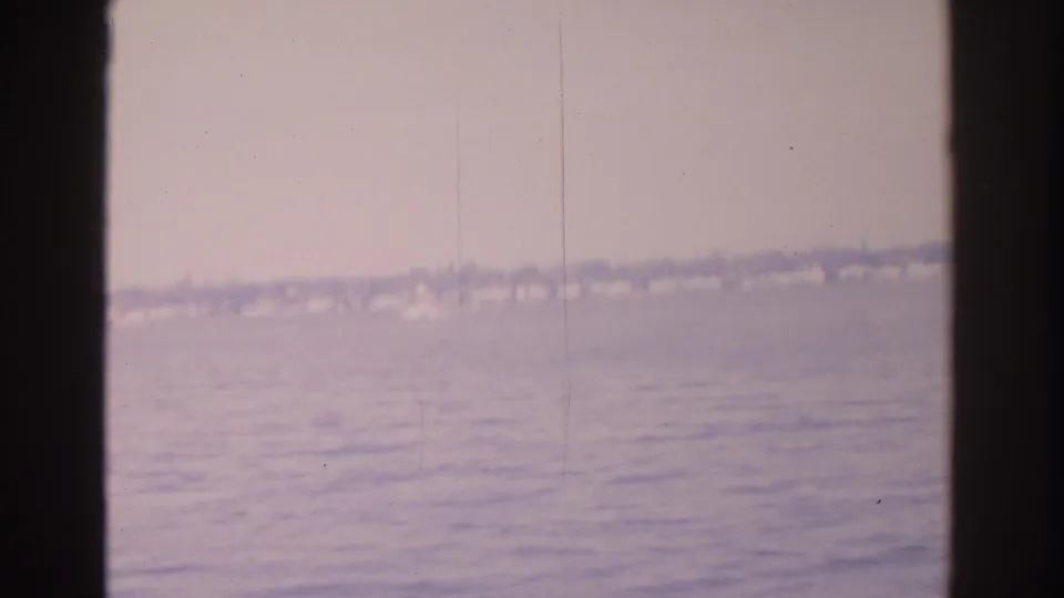 1962 Florida home-movie frame showing rippled water and a hazy distant shoreline with low homes and trees.