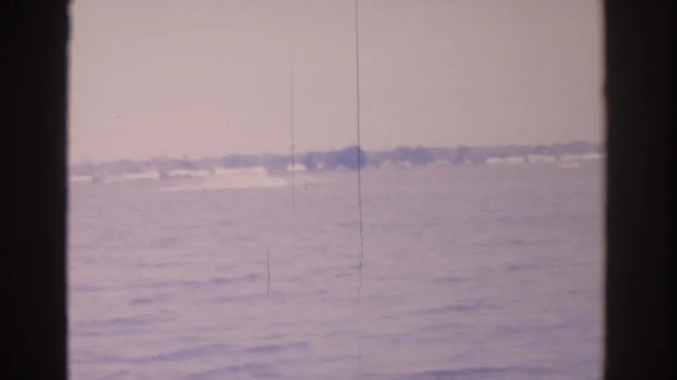 1962 8mm Florida bay view with hazy water, a distant low shoreline, faint wakes, and dark vertical frame borders.