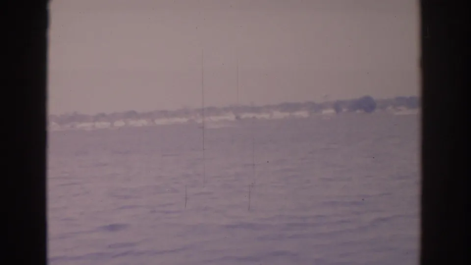 1962 Florida waterfront home-movie frame showing bay water, a far shoreline, film scratches, and a dark central border from the contact sheet.