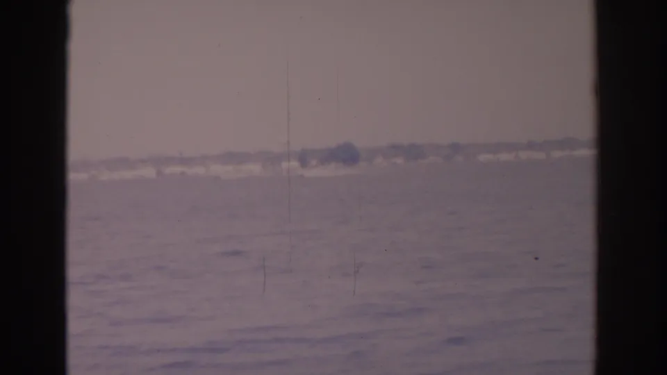 1962 8mm Florida bay frame showing calm water, a hazy far shoreline, faint wake texture, vertical scratches, and dark film borders.