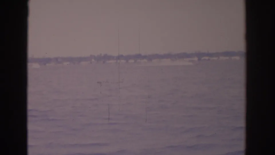 A 1962 Florida home-movie frame shows calm water and a hazy coastal strip, with dark vertical edges suggesting filming through an opening.