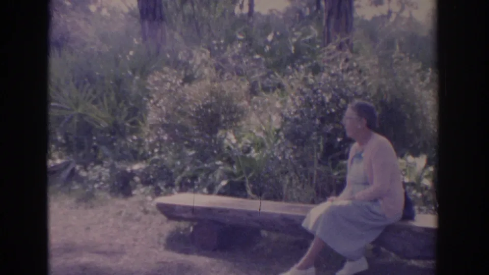 1962 Florida home-movie frame of an adult woman seated in profile on a stone bench, surrounded by bright garden foliage.
