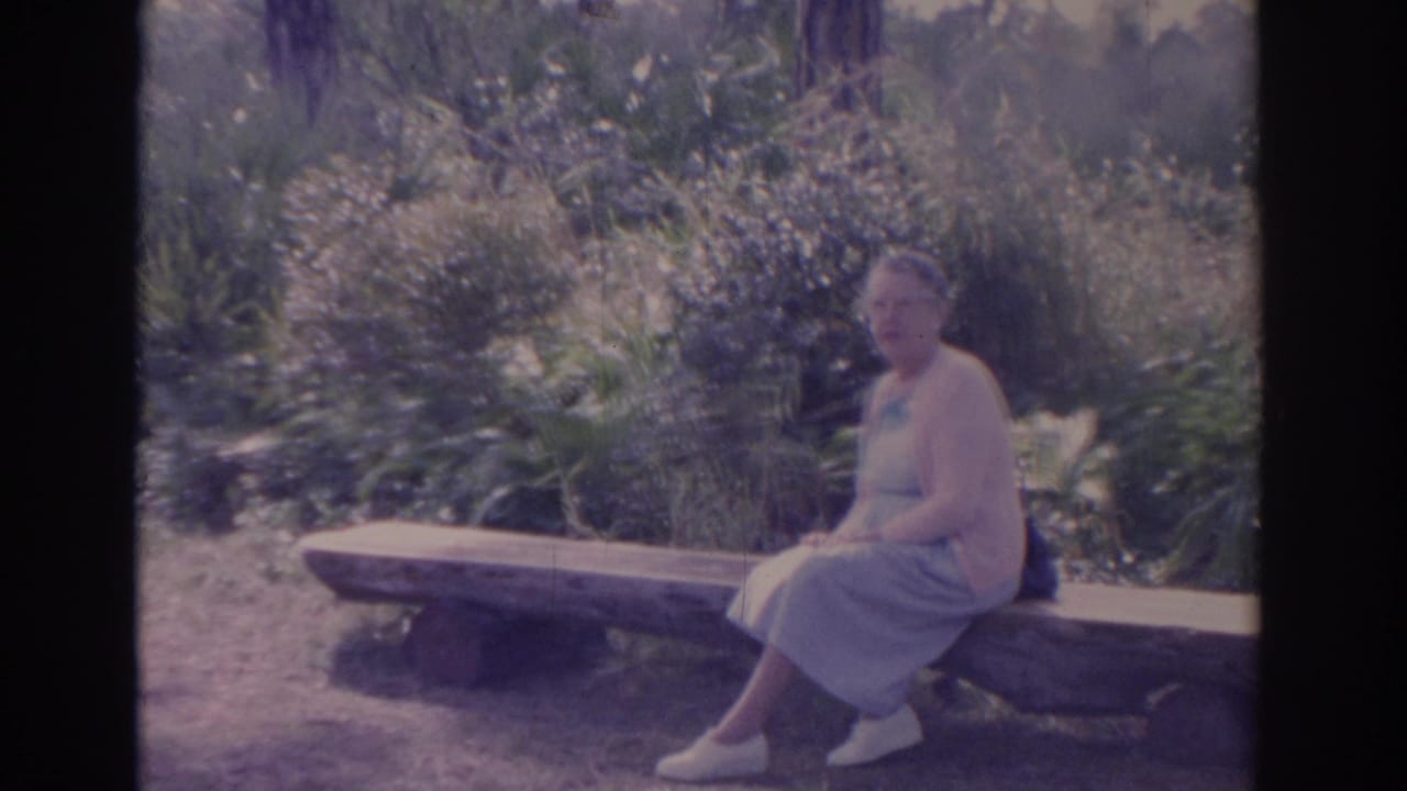Older woman in 1962 Florida home-movie footage sits on a rustic bench among dense outdoor foliage.