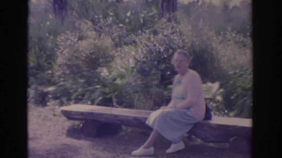 Older woman in 1962 Florida home-movie footage sits on a rustic bench among dense outdoor foliage.