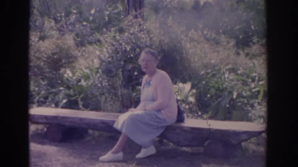 In a Florida garden setting, the woman remains seated on the bench with leafy plants and trees behind her.
