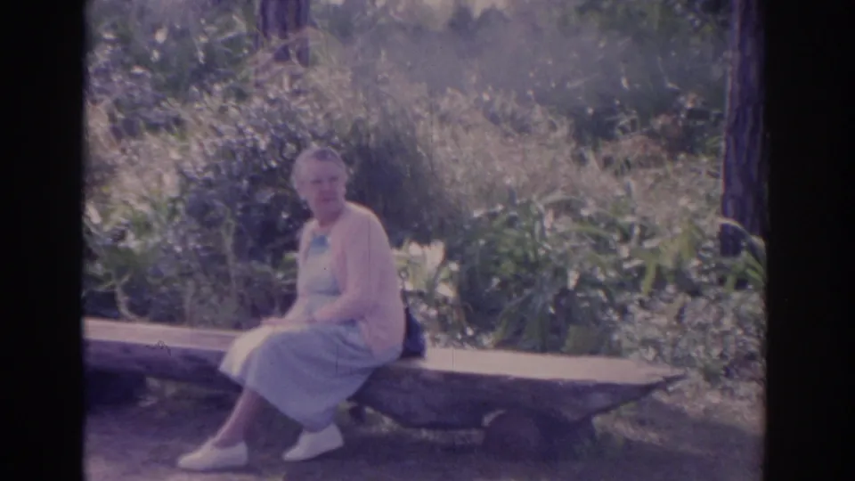 The 8mm frame shows the older woman seated near the end of a long bench, surrounded by Florida garden foliage.