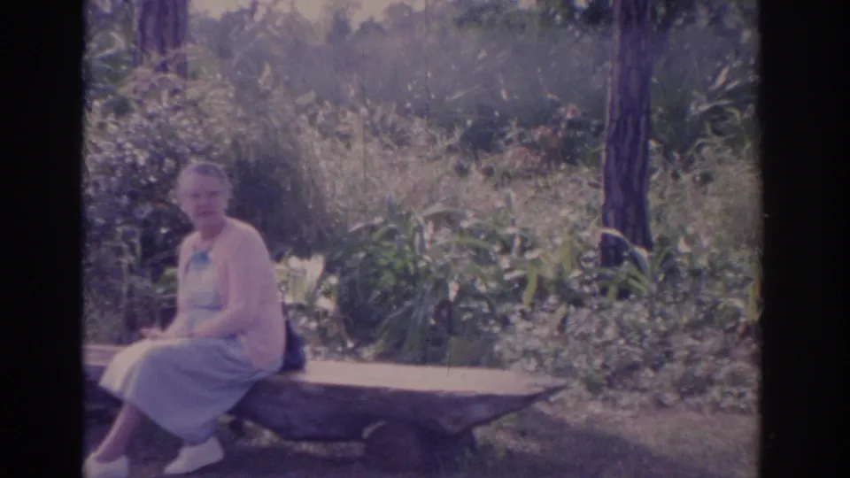 A soft 8mm frame shows the woman seated on the bench, with shrubs, grasses, and tree trunks filling the background.