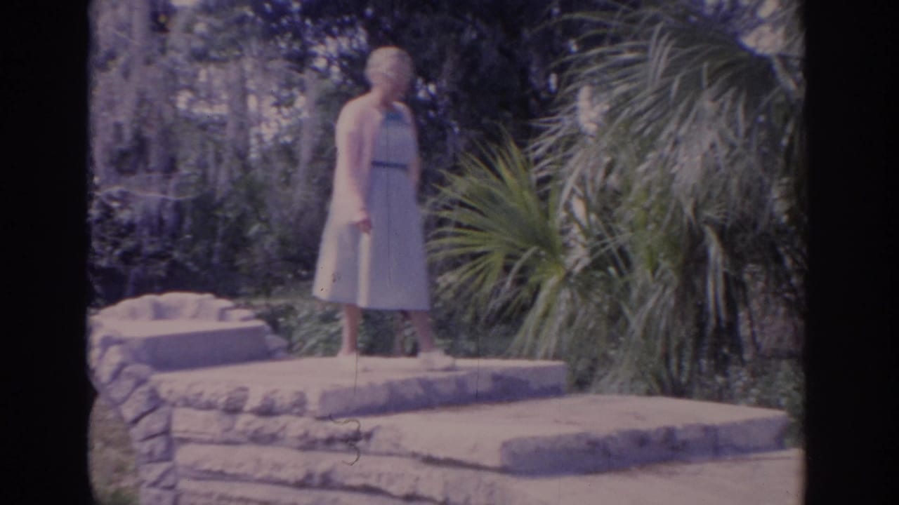 1962 Florida 8mm frame of an elderly woman in a light dress and cardigan standing on a rustic stone bridge area.