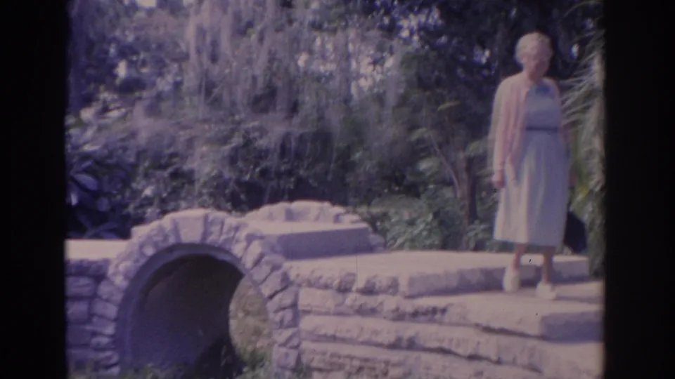 1962 Florida 8mm frame showing an elderly woman crossing a small arched stone bridge in a leafy garden.