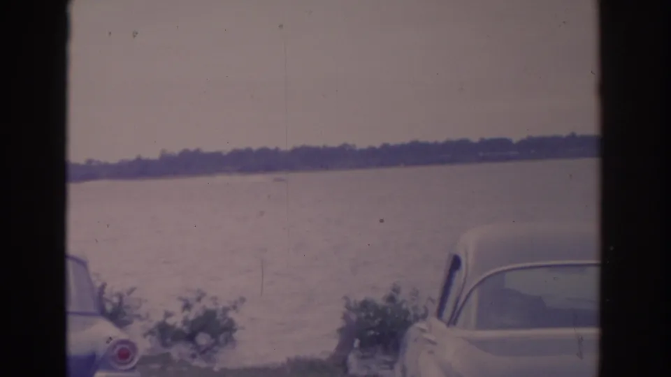 1962 Florida 8mm frame showing calm water, a distant tree-lined shore, shrubs, and early-1960s cars parked along the lakeside.