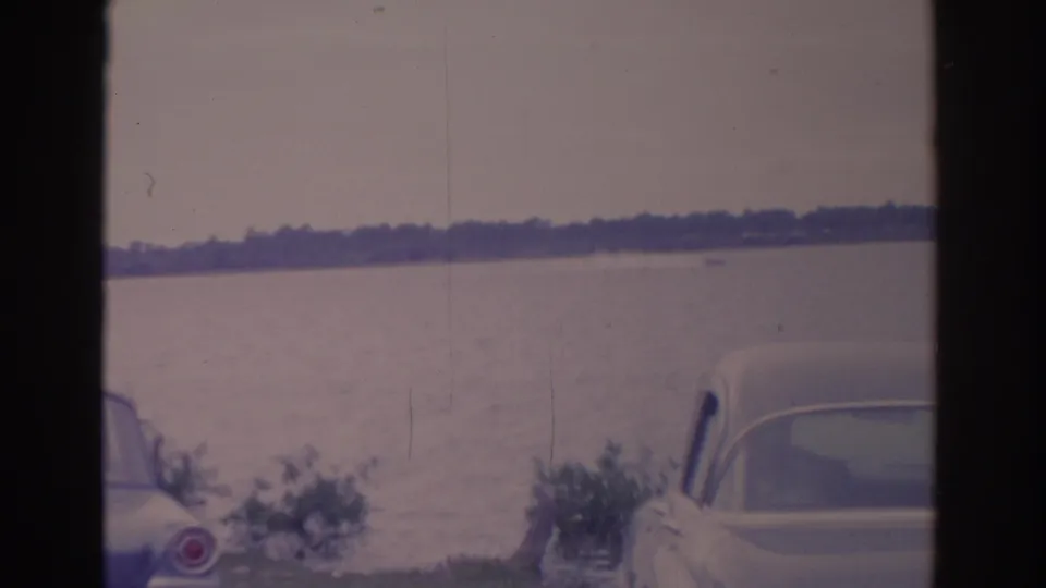 1962 Florida home-movie frame with a parked car beside the lake, distant shoreline trees, and a small boat moving across open water.