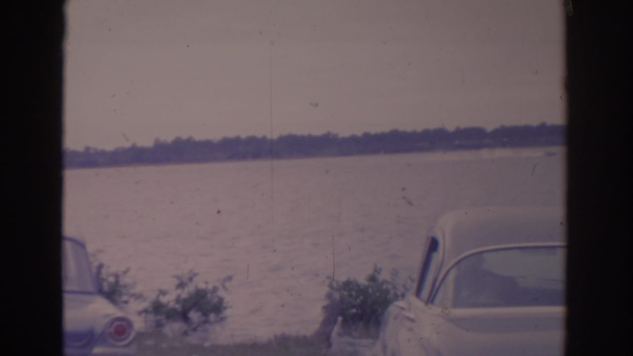 1962 Florida 8mm frame showing vintage cars partly cropped in the foreground beside a calm lake and hazy distant shoreline.