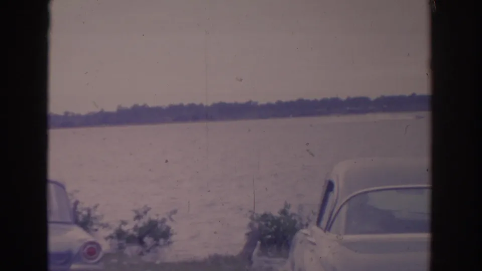 1962 Florida 8mm frame showing vintage cars partly cropped in the foreground beside a calm lake and hazy distant shoreline.