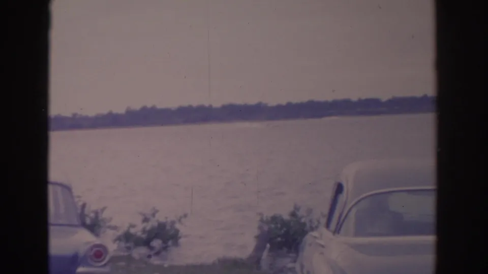 1962 Florida home-movie frame with parked early 1960s cars, lakeside shrubs, calm water, and a distant tree line.